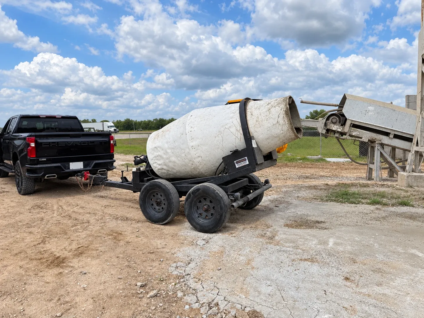 A black pickup hitched to a towable mixer under the batch plant's chute.