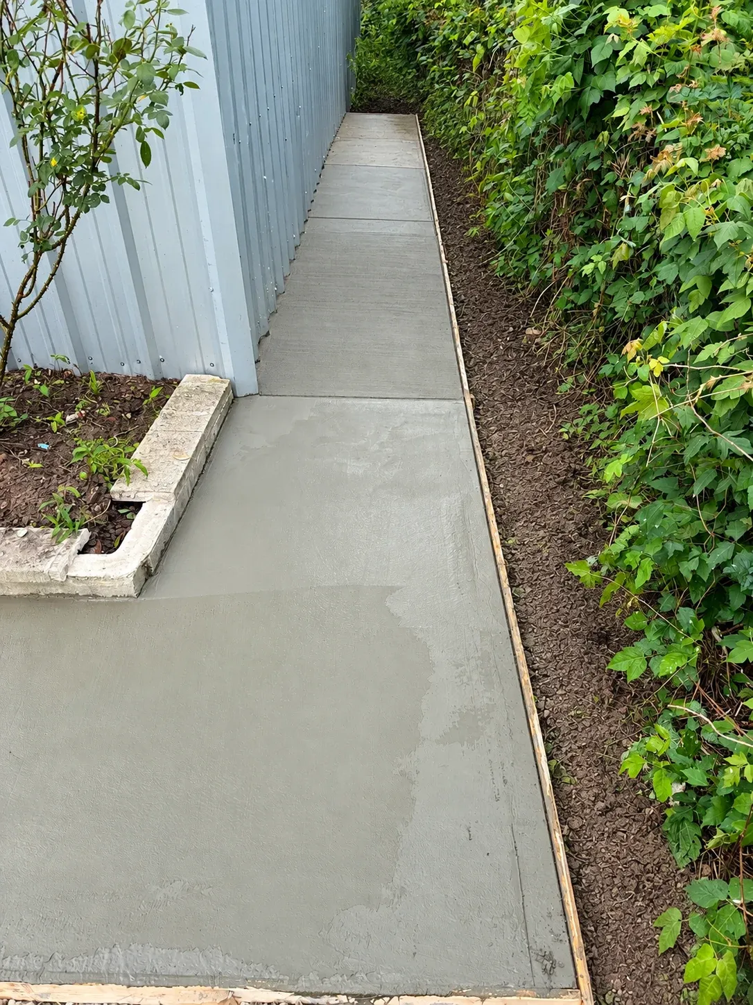 A fresh concrete walkway running beside a metal building, the forms still in place.