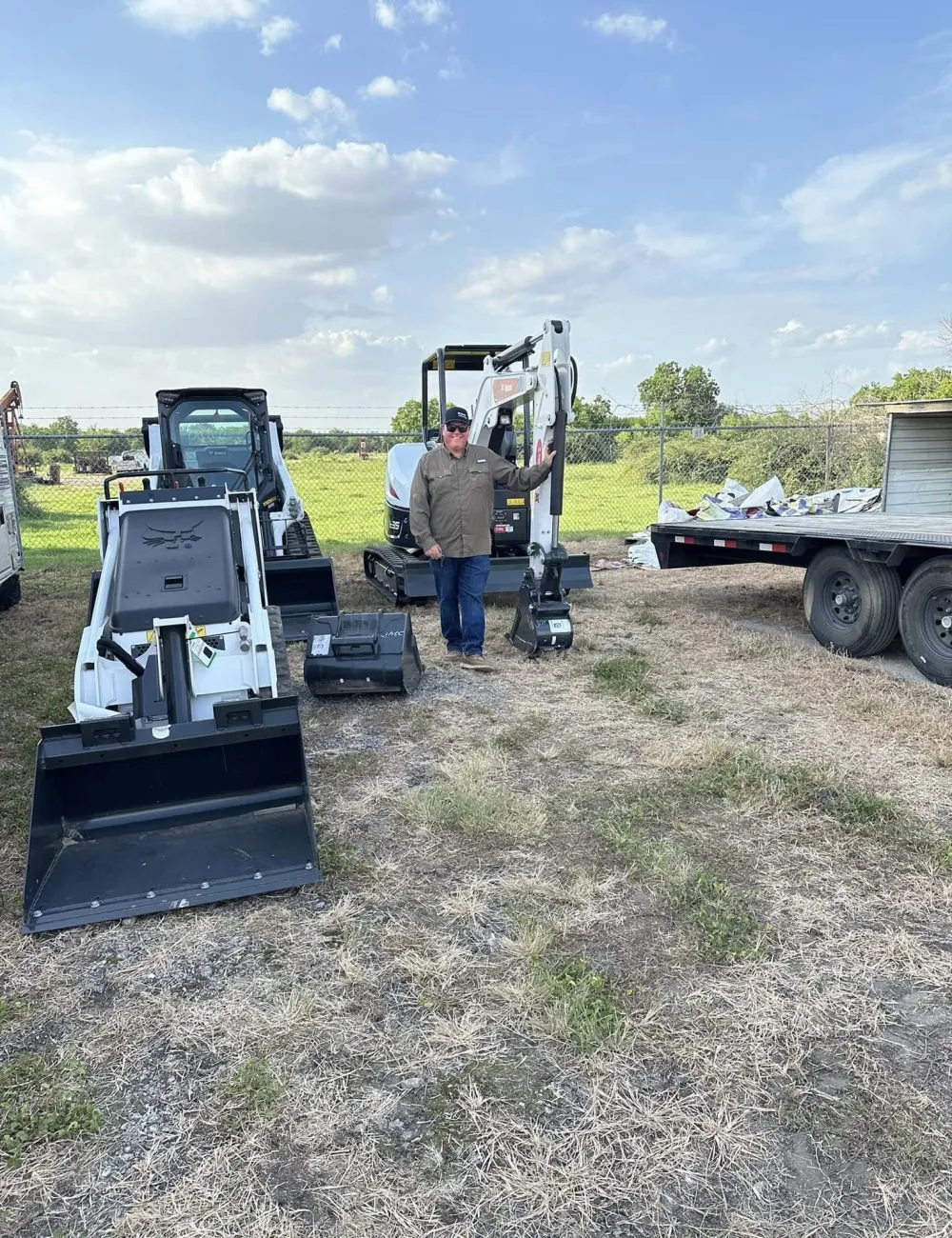John Melia standing beside a mini excavator and a skid steer at the yard.