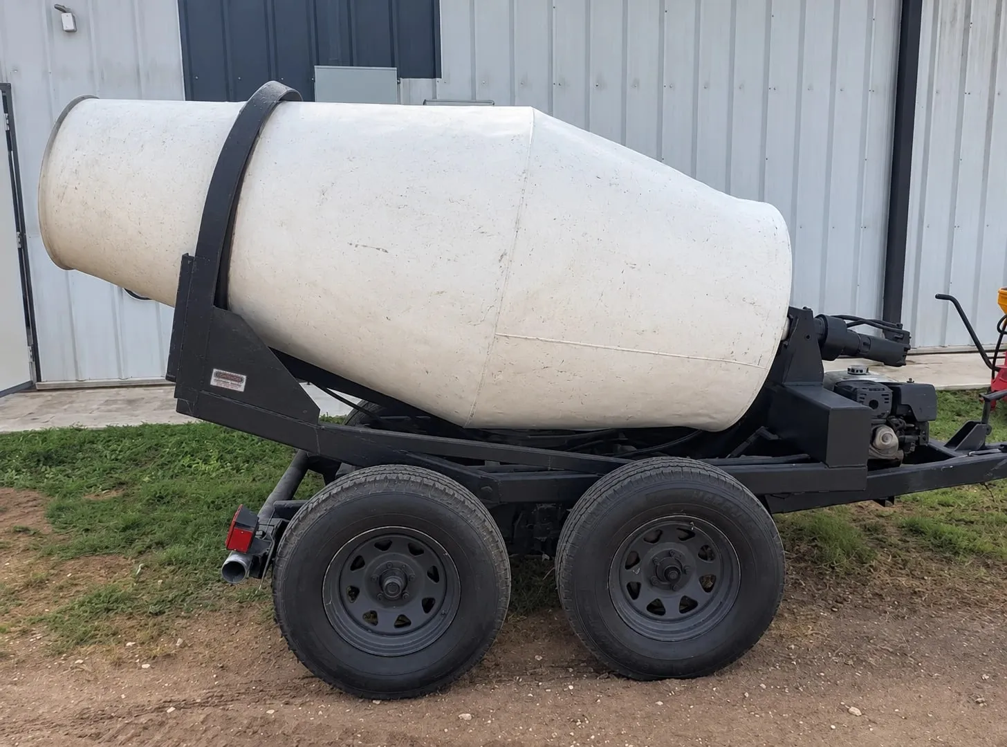A white towable concrete mixer on a black two-axle trailer, parked by a metal building.